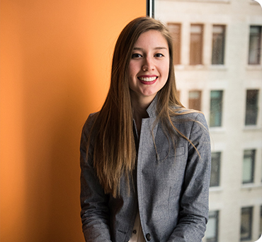 Young professional woman with long brown hair wearing a gray blazer smiling confidently in a modern office setting