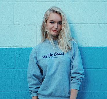 Young woman with long blonde hair wearing a gray Myrtle Beach sweatshirt standing against a blue two-tone wall