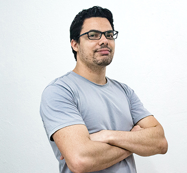 Confident man with glasses wearing a light gray t-shirt standing with arms crossed against a white background