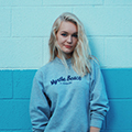 Young woman with blonde hair wearing a light grey sweatshirt standing against a blue gradient wall background