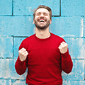 Man wearing a red sweater smiling and celebrating with fists clenched against a blue wall background
