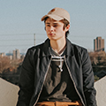 Young man wearing a cap and casual jacket standing outdoors with a cityscape in the background on a clear day