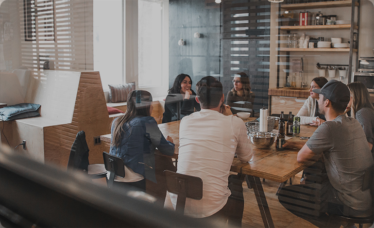 Group of young professionals having a casual meeting around a wooden table in a modern office with glass walls and wooden shelves.