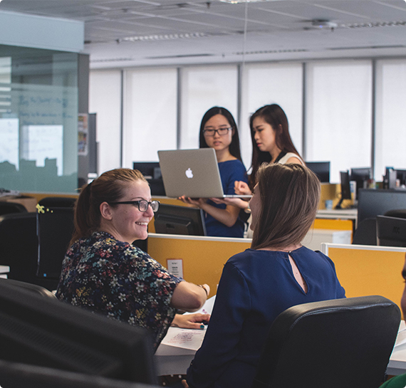 Four women collaborating in a modern office environment with computers, discussing work and sharing ideas.