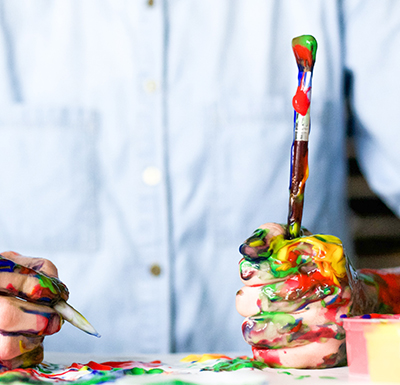 Close-up of hands covered in multicolored paint holding a paintbrush and pen while creating artwork on a table.