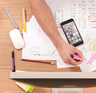 Person highlighting and annotating design sketches and notes with a smartphone and computer mouse nearby on a wooden desk.