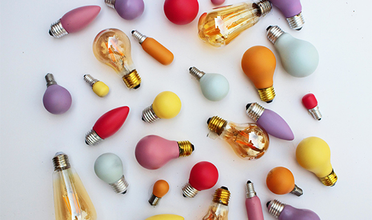 Various colorful and differently shaped light bulbs scattered on a white surface representing creativity and ideas.