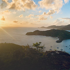 Sunset view over a coastal landscape with hills, calm sea, scattered boats, and partly cloudy sky.