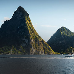 Tall volcanic mountains rise dramatically from the calm sea under a clear sky with sunlight casting shadows on the lush slopes.