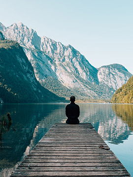 Person sitting on a wooden dock overlooking a calm lake surrounded by towering mountains under a clear sky