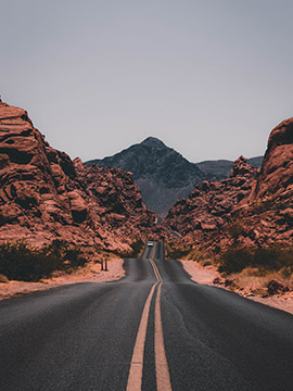 Two-lane road leading through red rocky desert terrain with mountains in the distance under clear sky