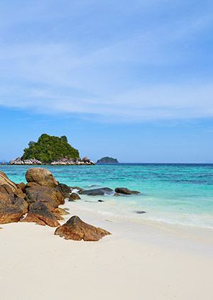 Sandy beach with large rocks in the foreground and a small lush island in the clear turquoise sea under a blue sky