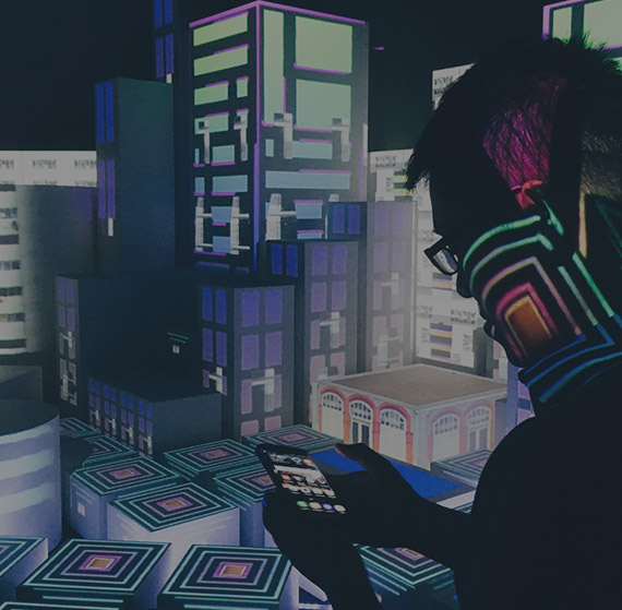 Person using a smartphone in a dark room with colorful geometric light projections on a miniature cityscape backdrop.