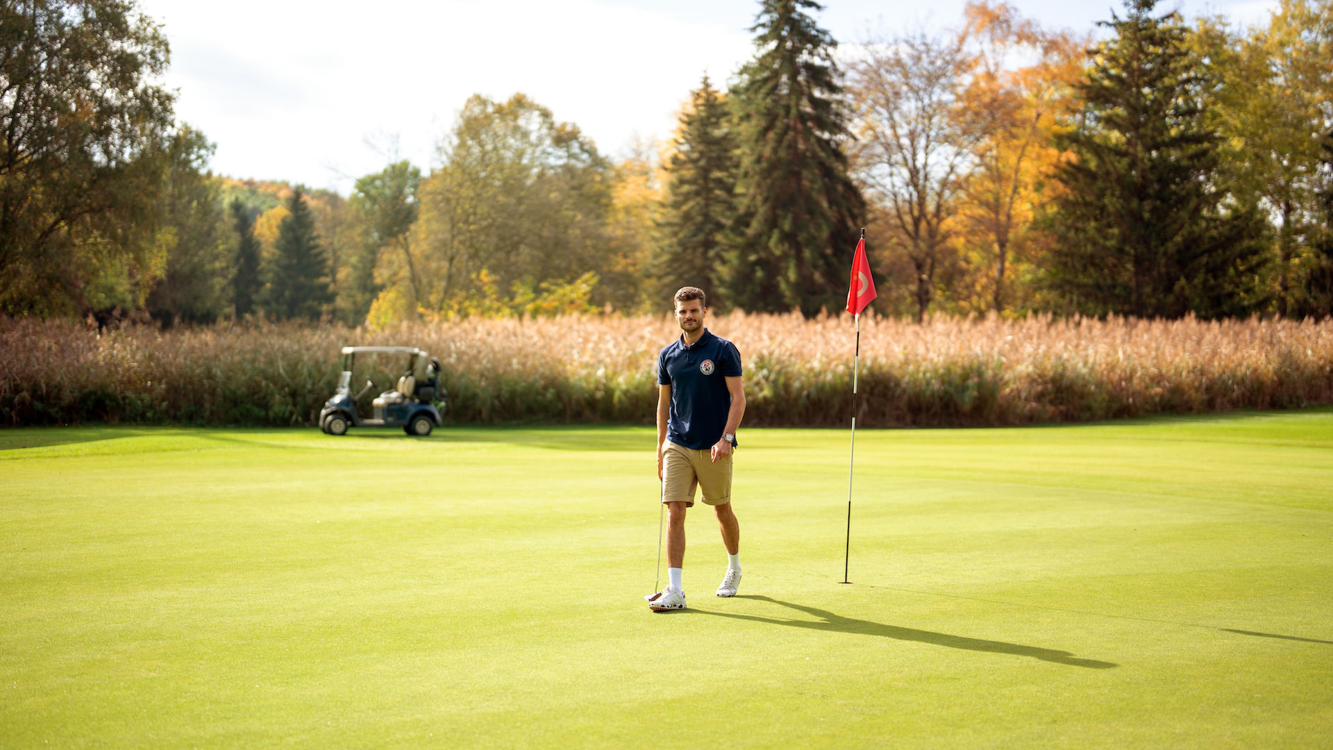 Man walking on a golf course near the hole flag with a putter in hand surrounded by autumn trees and a golf cart in the background
