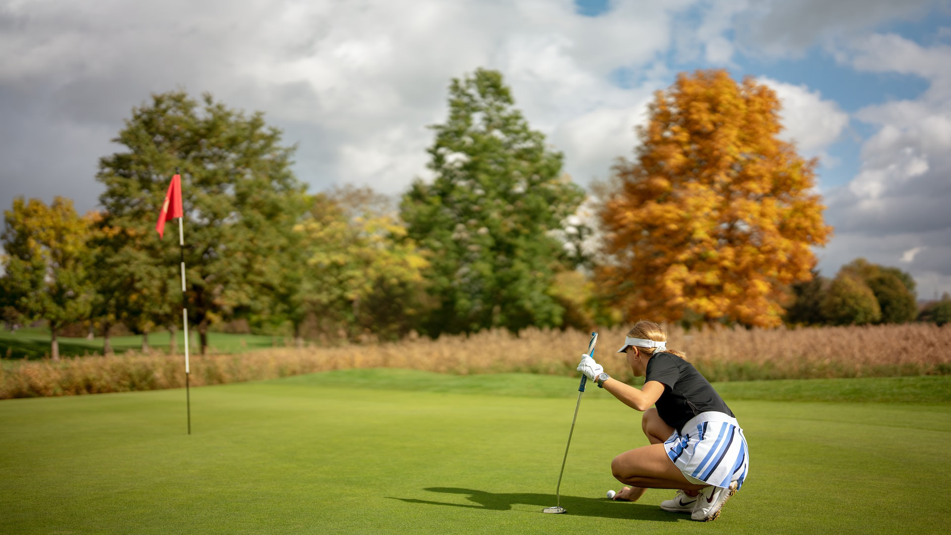 Female golfer in sports attire crouching on a putting green aiming to putt a golf ball near the hole with a red flag on a cloudy day.